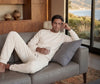 Man in a white outfit sitting on a couch in a modern living room with a scenic view.