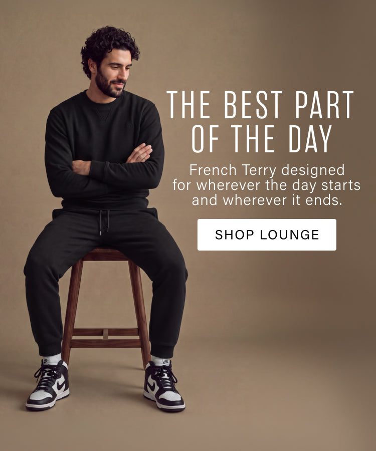 A man wearing a black long-sleeve shirt and black sweatpants sits on a wooden stool against a warm brown studio background. White text on the right reads 'THE BEST PART OF THE DAY' with a 'SHOP LOUNGE' button.